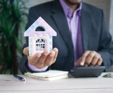 Businessman in suit holds model house, calculates real estate investments.